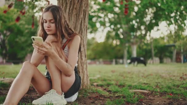 윗면에 아름다운 소녀는 의나무 아래푸른 잔디에 전화를 전화기에 사진들을 손가락으로 — 비디오