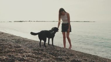 Beyaz bluzlu uzun dalgalı saçlı tatlı kız, çakıl taşı plajında siyah köpek deniz manzaralı oynuyor.