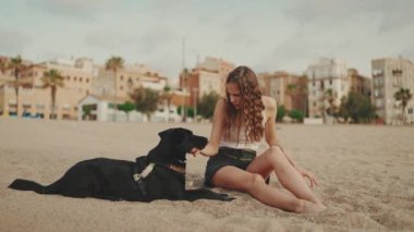Beyaz bluzlu uzun dalgalı saçlı güzel bir kız modern evdeki siyah köpekle kumda oturuyor. Kız evcil hayvanını okşuyor. Aşk ve sevgi