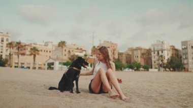 Beyaz saçlı, uzun dalgalı saçlı güzel bir kız modern ev arka planında siyah bir köpekle kumların üzerinde oturuyor. Kız evcil hayvanını eğitiyor. Sevgi ve sevgi