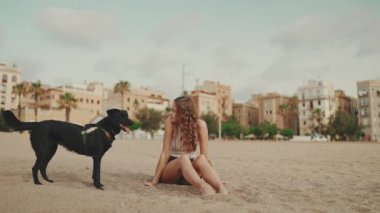Beyaz bluzlu uzun dalgalı saçlı güzel bir kız modern evdeki siyah köpekle kumda oturuyor. Kız evcil hayvanını okşuyor. Aşk ve sevgi