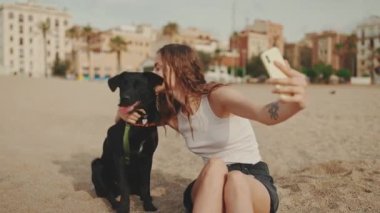 Beyaz bluzlu uzun dalgalı saçlı güzel kız modern bina arka planında siyah köpekle kumda otururken selfie çekiyor. Kız hayvanına sarılıyor.