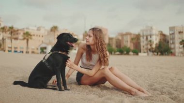 Beyaz bluzlu uzun dalgalı saçlı güzel bir kız modern bina arka planında siyah bir köpekle kumların üzerinde oturuyor. Kız evcil hayvanını okşuyor ve öpüyor