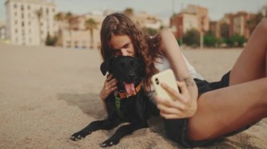 Beyaz bluzlu uzun dalgalı saçlı güzel kız modern bina arka planında siyah köpekle kumda otururken selfie çekiyor. Kız hayvanına sarılıyor.