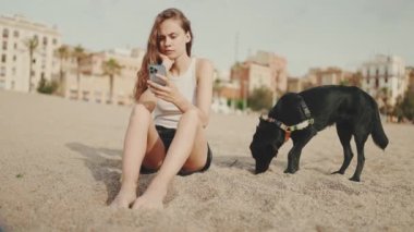 Elinde cep telefonu olan tatlı bir kız modern bina arka planında siyah bir köpekle kumsalda oturuyor. Güzel kız akıllı telefonuyla video ve fotoğraf izliyor.
