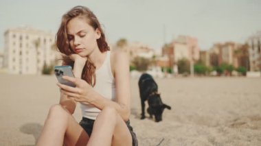 Elinde cep telefonu olan tatlı bir kız modern bina arka planında siyah bir köpekle kumsalda oturuyor. Güzel kız akıllı telefonuyla video ve fotoğraf izliyor.