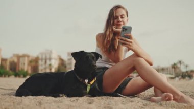 Elinde cep telefonu olan tatlı bir kız modern bina arka planında siyah bir köpekle kumsalda oturuyor. Güzel kız akıllı telefonuyla video ve fotoğraf izliyor.
