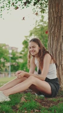 Kız, parkta evcil hayvanıyla birlikte çimlerin üzerinde, ağacın altında oturuyor. Köpeği çağır ve onu sev.