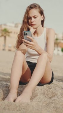 Elinde cep telefonu olan sevimli bir kız modern bina arka planında kumsalda oturuyor, kumsalda. Kız akıllı telefonuyla video ve fotoğraf izliyor.