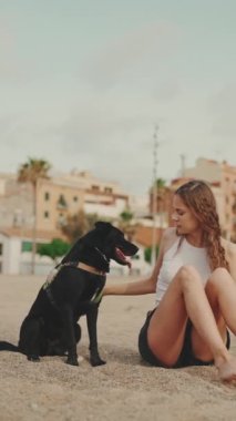 Beyaz saçlı, uzun dalgalı saçlı güzel bir kız modern ev arka planında siyah bir köpekle kumun üzerinde oturuyor. Kız evcil hayvanını eğitiyor. Sevgi ve sevgi