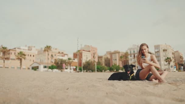 Cute Girl Telefonem Komórkowym Rękach Siedzi Piasku Plaży Czarnym Psem — Wideo stockowe