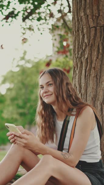 Vertical Video 漂亮的女孩 留着长长的波浪般的长发 头戴白色上衣 坐在公园里的树下绿草上 一边用手机 — 图库视频影像