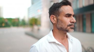 Yakın plan iş adamı sakallı, beyaz tişörtlü, olgun bir adam başını kameraya çeviriyor ve şehrin finans bölgesinde gülümsüyor.