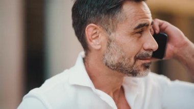 Yakın plan, sakallı, olgun iş adamı cep telefonu kullanıyor şehrin finans bölgesinde bankta oturuyor. Akıllı telefondan konuşan başarılı bir adam. Kamera hareket ediyor