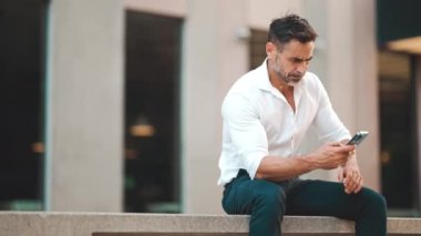 Beyaz gömlek giyen, sakallı, olgun bir iş adamı şehrin finans bölgesinde bankta oturuyor. Başarılı adam akıllı telefondan bilgi aktarıyor.