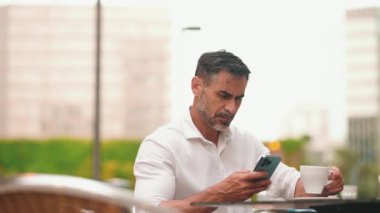 Sakallı, beyaz gömlekli, olgun bir işadamı cep telefonu kullanıyor. Kafe masasında oturan, kahve içen ve akıllı telefonda yazan başarılı bir adam.