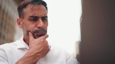 Düz sakallı, olgun bir iş adamı laptopta beyaz gömlek giyiyor bilgisayar açık kafede oturuyor. Doğada açık havada, kafede oturan başarılı bir adam. Mobil ofis serbest.