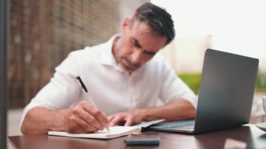 Sakallı olgun bir iş adamı, dizüstü bilgisayarlı bir kullanıcı ve açık kafede oturup notlar alıyor. Doğa Mobil Ofis Freelance 'da açık kafedeki masada oturan adam. Kamera eline odaklan