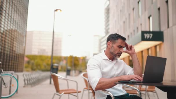 成熟的商人留着整洁的胡子 穿着白色衬衫 坐在笔记本电脑上 坐在室外咖啡馆里 一个成功的男人坐在野外的咖啡馆桌旁 在大自然中自由活动 — 图库视频影像