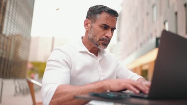 在笔记本电脑上有胡子用户的老实人商人坐在外面的咖啡馆里 一个成功的男人坐在户外的咖啡馆桌旁 坐在自然移动办公室的自由职业上 成功的交易 — 图库视频影像