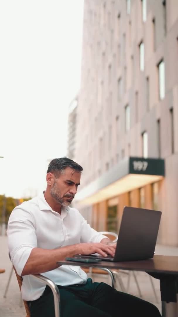 垂直视频 在笔记本电脑上有整齐胡子用户的成熟商人坐在室外咖啡馆里 一个成功的男人坐在野外的咖啡馆桌旁 在大自然中自由活动 — 图库视频影像
