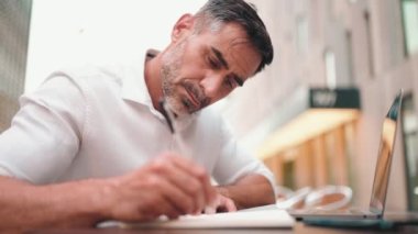 Sakallı olgun bir iş adamı, dizüstü bilgisayarlı bir kullanıcı ve açık kafede oturup notlar alıyor. Doğada açık havada, kafede oturan başarılı bir adam. Mobil ofis serbest.