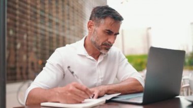 Sakallı olgun bir iş adamı, dizüstü bilgisayarlı bir kullanıcı ve açık kafede oturup notlar alıyor. Doğada açık havada, kafede oturan başarılı bir adam. Mobil ofis serbest.