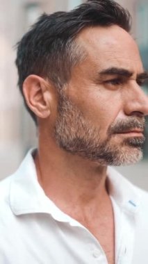 Yakın plan iş adamı sakallı, beyaz tişörtlü, olgun bir adam başını kameraya çeviriyor ve şehrin finans bölgesinde gülümsüyor.