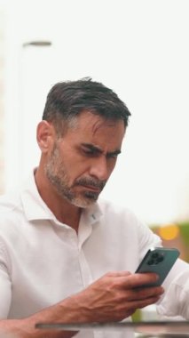 Sakallı, beyaz gömlekli, olgun bir işadamı cep telefonu kullanıyor. Kafe masasında oturan, kahve içen ve akıllı telefonda yazan başarılı bir adam.