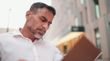 Beyaz gömlekli, dokümanlara bakan olgun iş adamı açık kafede oturuyor. Doğada açık havada, kafede oturan başarılı bir adam. Mobil ofis serbest.