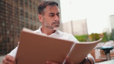 Beyaz gömlekli, dokümanlara bakan olgun iş adamı açık kafede oturuyor. Doğada açık havada, kafede oturan başarılı bir adam. Mobil ofis serbest.