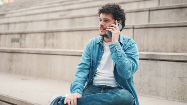 Kot gömlekli genç sakallı adam yüksek basamaklarda oturuyor ve cep telefonu kullanıyor. Akıllı telefondan konuşan adam.