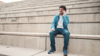 Kot gömlekli genç sakallı adam yüksek basamaklarda oturuyor ve cep telefonu kullanıyor. Akıllı telefondan konuşan adam.