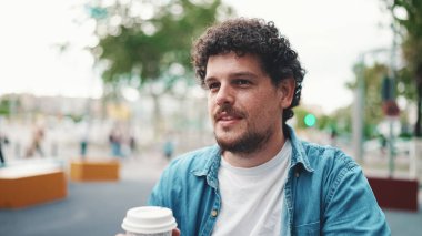 Kot gömlekli, kablosuz kulaklıklı ve modern şehir arka planında kahve içen genç sakallı adamın yakın plan portresi. Kahve içen adamın profili.