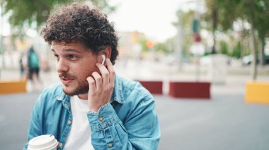 Kot gömlekli, kablosuz kulaklıklı ve modern şehir arka planında kahve içen genç sakallı adamın yakın plan portresi. Kahve içen adamın profili.