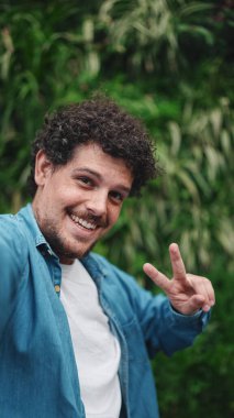 Yakın plan, genç sakallı adam akış yapıyor, yeşil yapraklı arka planda video çağrısı var.