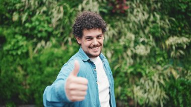 Kotlu tişörtlü genç sakallı adamın yakın çekimi. Sınıfını gösteriyor ve modern şehir arka planında kameraya gülüyor.