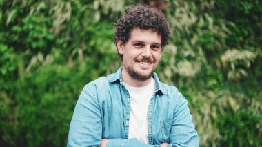 Yakın plan, kot gömlekli genç sakallı adam elini uzatıyor ve modern şehrin arka planında kameraya gülümsüyor.