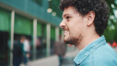 Kot gömlekli genç sakallı adam modern şehir arka planında yaya geçidinden geçerken gülümsüyor.