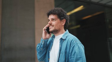Kot pantolon giyen genç sakallı adam cep telefonuyla konuşuyor ve modern binalarda yürüyor.