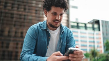 Kot gömlekli gülümseyen genç sakallı adam cep telefonu kullanıyor. Modern şehir geçmişinde akıllı telefon sosyal ağlarına bakan bir adam.