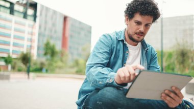 Kot gömlekli genç sakallı adam modern binalarda oturuyor ve tablet kullanıyor.