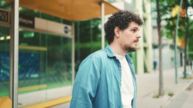 Yakın plan, kot gömlekli genç sakallı adam otobüs durağında duruyor ve modern şehir arka planında şehir ulaşımını bekliyor.