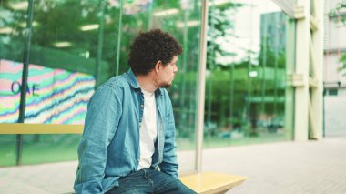 Kot pantolon giyen genç sakallı adam otobüs durağında oturur şehir taşıma saatlerini öğrenir ve modern şehir arka planında otobüs beklerken başka yere bakar.