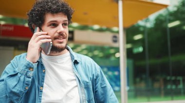 Kot gömlekli genç adam otobüs durağında duruyor otobüs bekliyor ve modern şehir arka planında cep telefonuyla konuşuyor.