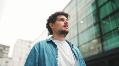 Yakın plan, kot gömlekli adam sokakta yürüyor, duruyor ve modern binaların üst katlarına bakıyor.