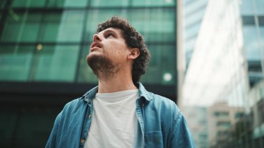 Yakın plan, kot gömlekli adam sokakta yürüyor, duruyor ve modern binaların üst katlarına bakıyor.