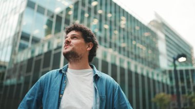 Yakın plan, kot gömlekli adam sokakta yürüyor, duruyor ve modern binaların üst katlarına bakıyor.