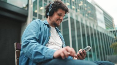 Kot gömlekli, kulaklıklı, bankta oturan, müzik dinleyen ve modern binalarda cep telefonu kullanan genç sakallı adamın yakın çekimi.