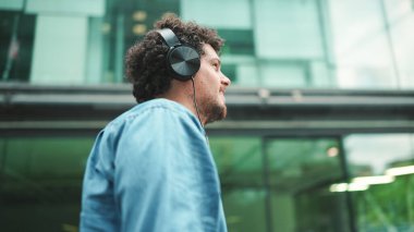 Kot gömlekli duygusal genç adam kulaklıkla sokakta yürüyor müzik dinliyor ve şarkı söylüyor, elinde cep telefonu ile modern şehir manzarası arka planında.